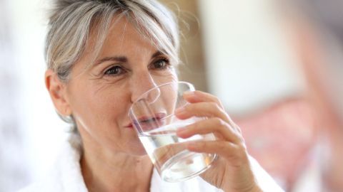 Eine Frau trinkt ein Glas Wasser, um ihre trockene Haut zu hydratisieren, in einem hellen, freundlichen Wohnraum.
