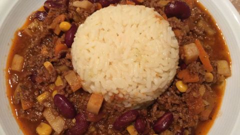 Ein Teller Chili con Carne zeigt Fleisch und Bohnen in einer würzigen Sauce, umgeben von beigefarbenem Reis.