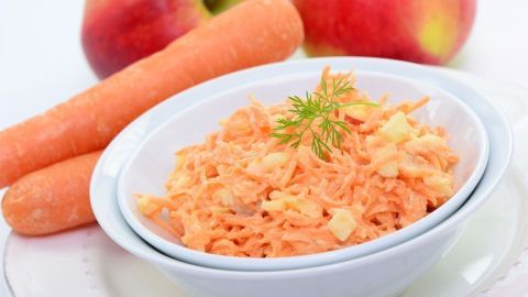 Ein frischer Karotten-Apfel-Meerrettich-Salat liegt in einer Schüssel, umgeben von Äpfeln und Karotten auf einem weißen Tisch.
