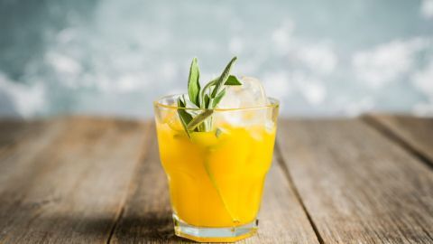 Ein Glas mit einer gelben Tequila-Mischung steht auf einem Holztisch, dekoriert mit einem Grün und Eis.