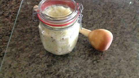 Ein Glas mit frisch gemachtem Sauerkraut steht auf einer glatten Oberfläche neben einem Holzlöffel in einer Küchenumgebung.
