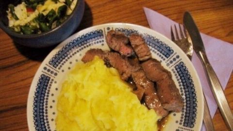 Ein saftiges T-Bone-Steak liegt neben cremigem Kartoffelpüree auf einem Teller, umgeben von einem Salat.