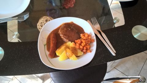 Ein saftiger Schweinebraten liegt auf einem weißen Teller, umgeben von Kartoffeln und Karotten, auf einem eleganten Tisch.