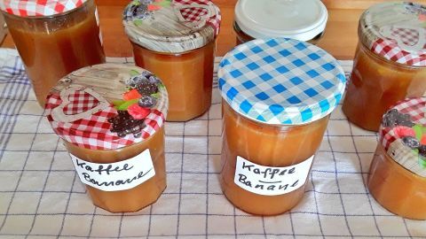 Gläser mit Kaffee-Bananen-Orangen Marmelade stehen auf einem Tisch, umgeben von karierten Deckeln und einer Gittermatte.