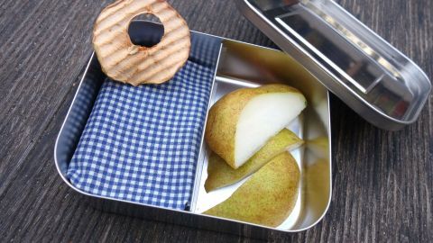Eine Edelstahlbox mit einer Wachstuch-Abdeckung, gefüllt mit Birnenscheiben und einem Holzspielzeug, auf einem Tisch.