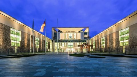 Ein modernes Gebäude mit Glasfassade steht im Zentrum, umgeben von einem klaren Abendhimmel und einer gepflasterten Fläche.