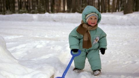 Ein Kleinkind in einem warmen Anzug spielt im Schnee und schaufelt mit einem blauen Schaufel im winterlichen Wald.