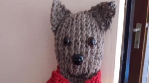 Ein gestrickter Teddy sitzt auf einem blauen Untergrund, trägt ein gelbes Oberteil und eine rote Schalsocke.