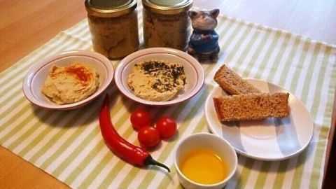 Zwei Schalen mit Hummus und frischem Gemüse liegen auf einem Tisch, umgeben von Kichererbsengläsern und Brot.