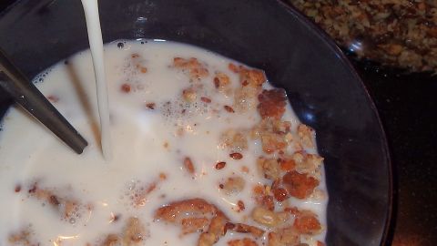 Eine Schüssel mit Knuspermüsli wird mit Milch gefüllt, während eine große Tüte mit Müsli im Hintergrund zu sehen ist.