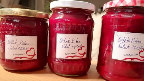 Drei Gläser mit roten Rüben im Glas, frisch konserviert und auf einem Holzstamm platziert. Labels beschreiben den Inhalt.
