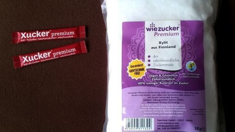 Ein Beutel mit zuckerfreiem Süßstoff liegt neben zwei Verpackungen mit dem Aufdruck „Xucker“. Ideal für Kariesprophylaxe.