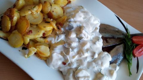 Geröstete Kartoffeln liegen neben frischen Matjesfilets, die in einer cremigen Sauce serviert werden, angerichtet auf einem weißen Teller.