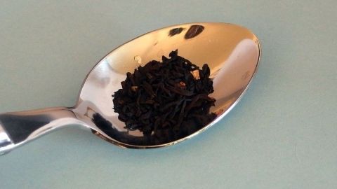 Gegen Knoblauchfahne hilft es 1 Minute lang schwarzen Tee zu kauen. Ein metallischer Löffel hält eine kleine Menge schwarzer, zerbrochener Knoblauchfahne auf einer hellen Unterlage.
