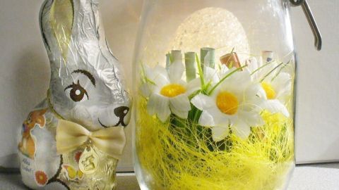 Ein dekoratives Glas mit Blumen und grünem Gras steht neben einem Schokoladenhasen, perfekt für ein Geldgeschenk zu Ostern.