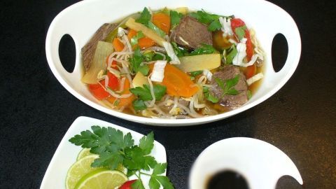 Eine Schüssel asiatische Rindfleisch-Gemüsesuppe steht auf einem Tisch, umgeben von frischen Kräutern und Limetten.