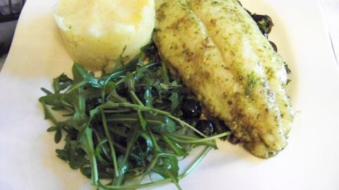 Ein gegrillter Fisch liegt auf einem Bett aus Oliven, garniert mit frischem Rucola und cremigem Kartoffelpüree.