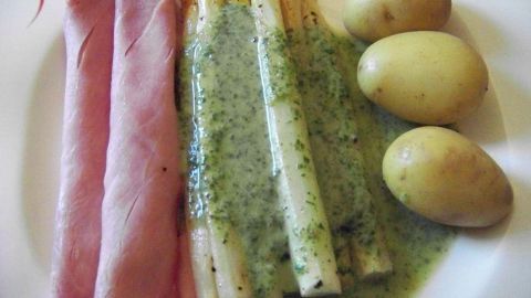 Drei gekochte Kartoffeln liegen neben gebackenem Spargel, der mit Kräutersoße und einem Stück Schinken serviert wird.