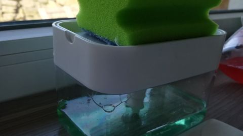 Ein Spülmittelspender steht auf einem Tisch und hält einen Schwamm, während eine helle Umgebung sichtbar ist.