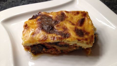 Ein Stück Moussaka liegt auf einem weißen Teller, leicht gebräunt und appetitlich, umgeben von einem dunklen Hintergrund.