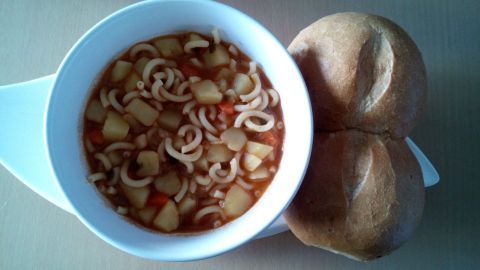 Eine Schüssel Kartoffel-Nudel-Suppe mit gemischtem Gemüse steht auf einem Tisch, begleitet von zwei Brötchen.