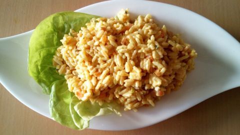 Ein Reissalat mit Curry liegt auf einem weißen Teller, umgeben von einem Blatt frischem Gemüse und präsentiert eine appetitliche, gelbe Farbe.