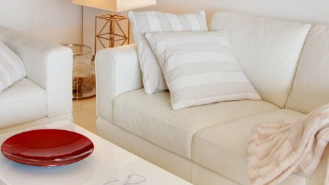 Eine weiße Ledercouch mit Kissen liegt im hellen Raum, während ein roter Teller auf dem Tisch steht.
