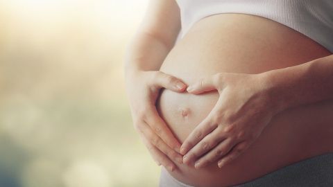 Ein schwangere Frau formt mit ihren Händen ein Herz auf ihrem Bauch, umgeben von weichem, verschwommenem Licht.