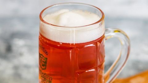 Ein gefüllter Glasbecher mit schäumendem Getränk steht auf einem Holzbrett, umgeben von Eiswürfeln in einem neutralen Raum.