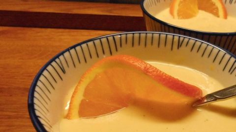 Ein schaumiger Orangenpudding in einer Schale, dekoriert mit einer Orangenscheibe, steht auf einem Holzbrett.
