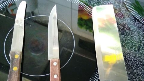 Drei scharfe Messer mit Holzgriffen liegen auf einer schwarzen Herdplatte, bereit für das Schneiden von Zutaten.