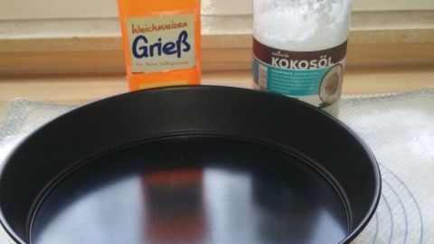 Eine schwarze Backform steht auf einer Arbeitsplatte, umgeben von Kokosöl und Grieß, bereit zum Backen.