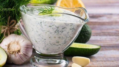 Ein Glas Knoblauchdressing aus Kefir steht auf einem Holztisch, umgeben von frischem Gemüse und Kräutern.