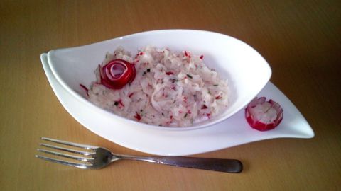 Ein frischer Rettich-Radieschen-Salat liegt in einer eleganten, weißen Schale auf einem Holztisch, umgeben von einem Besteck.