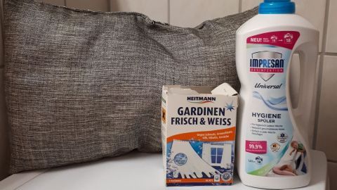 Ein graues Kissen liegt auf einer weißen Oberfläche, daneben steht ein Waschmittel und eine Packung für Gardinen.