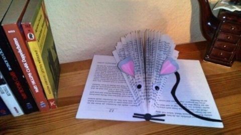 Ein gefaltetes Buch in Form einer Maus liegt auf einem Blatt Papier, umgeben von bunten Bücherregalen im Hintergrund.