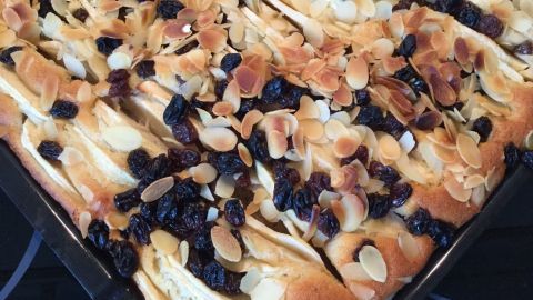 Ein goldbrauner Apfel-Blechkuchen liegt auf einem Blech, garniert mit Mandelsplittern und Rosinen in einer Küchenumgebung.