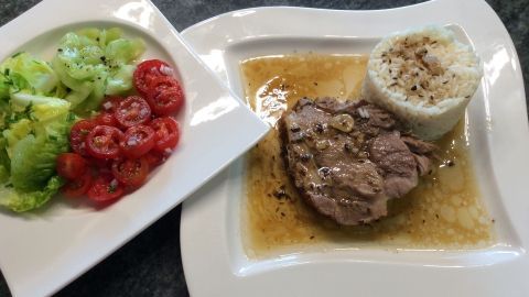 Ein saftiger Schweinsbraten liegt auf einem Teller mit Soße, begleitet von Reis und einem frischen Salat.