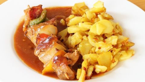 Saftige andalusische Spieße liegen auf einem Teller, umgeben von knusprig gebratenen Kartoffeln in einer aromatischen Soße.