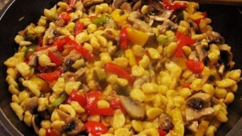 Eine bunte Mischung aus Knöpfle, Gemüse und Schweinegeschnetzeltem wird in einer Pfanne gebraten, umgeben von einer Küche.