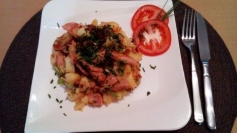 Auf einem weißen Teller liegt eine gut belegte Lyonerpfanne mit Fleisch, Zwiebeln und Kräutern, umgeben von Tomatenscheiben.