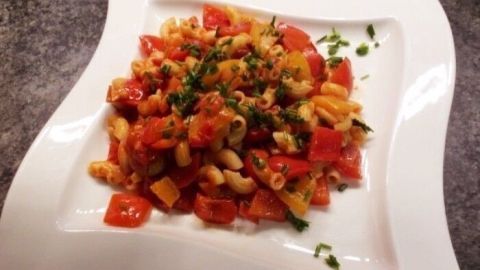 Ein farbenfrohes Letscho-Gericht auf einem weißen Teller, bestehend aus Nudeln, Tomaten und Kräutern in einer rustikalen Küche.
