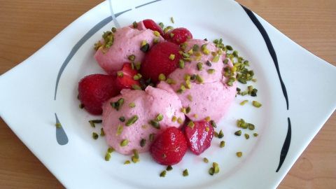 Auf einem weißen Teller liegt eine Erdbeer-Mousse mit frischen Erdbeeren und gehackten Pistazien, schön angerichtet.