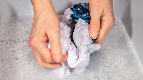 Hände waschen ein gebügelfreies T-Shirt in seifigem Wasser in einem Waschbecken, um Flecken zu entfernen.