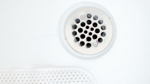 Ein Ablaufsieb in einer weißen Badewanne oder Dusche, umgeben von einer strukturierten Mattenoberfläche, zeigt Haarreste.