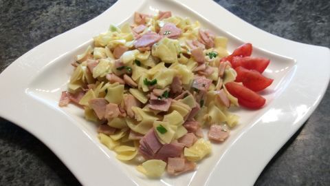 Ein Teller mit Schinkenfleckerln ist schön angerichtet, während bunte Tomatenscheiben daneben liegen.