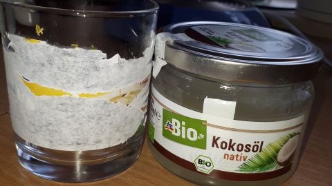 Ein Glas mit ablösenden Etiketten steht neben einem offenen Glas Kokosöl auf einem Holztisch in einer hellen Umgebung.