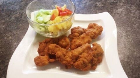 Paniertes Hühnerfleisch liegt auf einem weißen Teller neben einer bunten Gartensalat-Schüssel auf einer dunklen Arbeitsfläche.