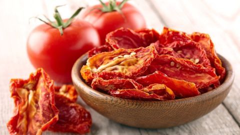 Getrocknete Tomaten liegen in einer Schüssel aus Holz, umgeben von frischen, roten Tomaten und einem Holzuntergrund.