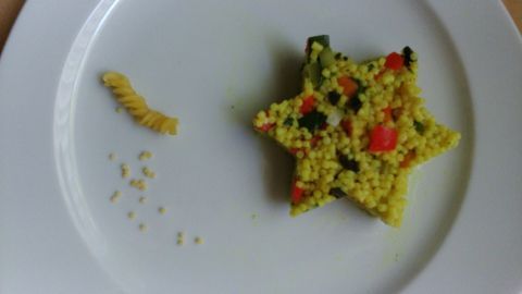 Ein bunter Graupennudelsalat hat die Form eines Sterns und liegt auf einem weißen Teller, neben einer kleinen Pasta.